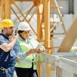 Two construction workers in hardhats discuss plans using a tablet and walkie talkie on-site.
