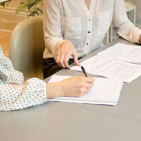 woman signing on white printer paper beside woman about to touch the documents